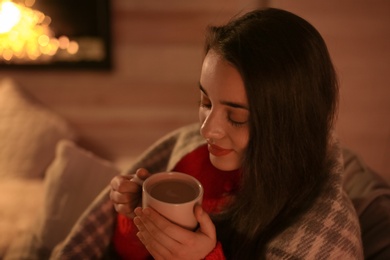 Photo of Young woman drinking coffee near decorative fireplace at home. Winter season