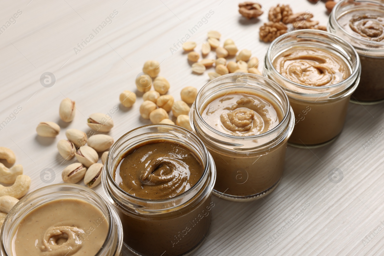 Photo of Many tasty nut butters in jars and nuts on white wooden table, closeup