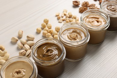 Many tasty nut butters in jars and nuts on white wooden table, closeup