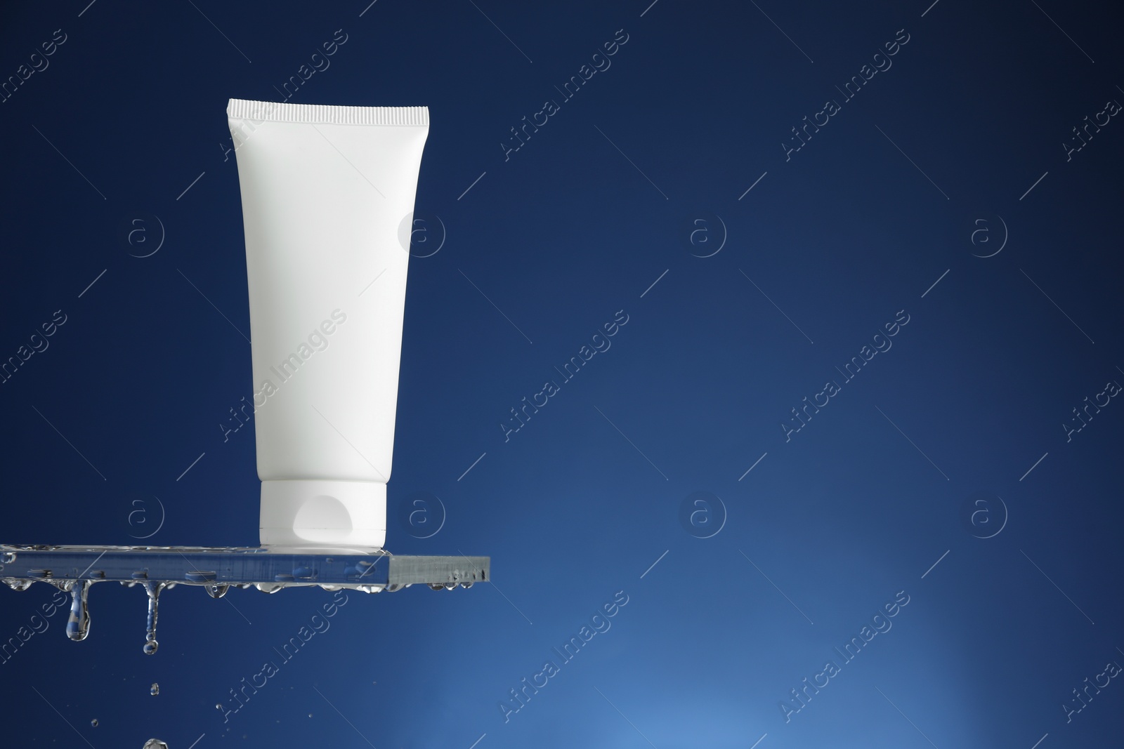 Photo of Moisturizing cream in tube on glass with water drops against blue background. Space for text