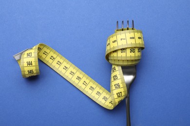 Fork with measuring tape on blue background, top view. Diet concept