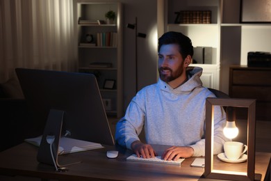 Photo of Home workplace. Happy man working with computer at wooden desk in room at night