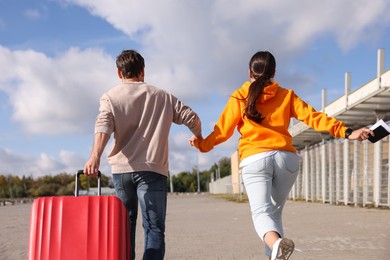 Photo of Being late. Couple with red suitcase running outdoors, back view
