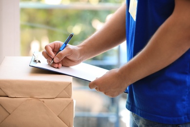 Photo of Young courier with parcels and clipboard against blurred background, closeup. Delivery service