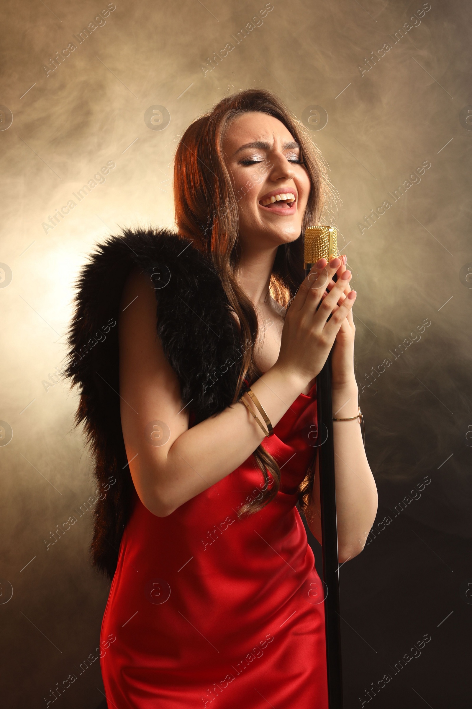 Photo of Beautiful young woman in stylish dress with microphone singing on dark background in color lights and smoke