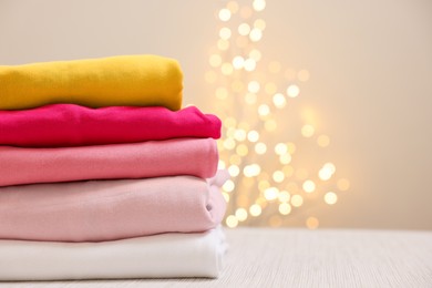 Photo of Stack of folded clothes on wooden table against beige background with blurred lights, space for text