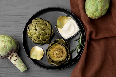 Delicious cooked artichokes with tasty sauce served on grey wooden table, flat lay