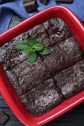 Delicious chocolate brownie with mint in baking dish on black wooden table, flat lay