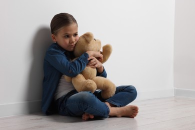 Photo of Child abuse. Upset girl with toy sitting on floor near white wall