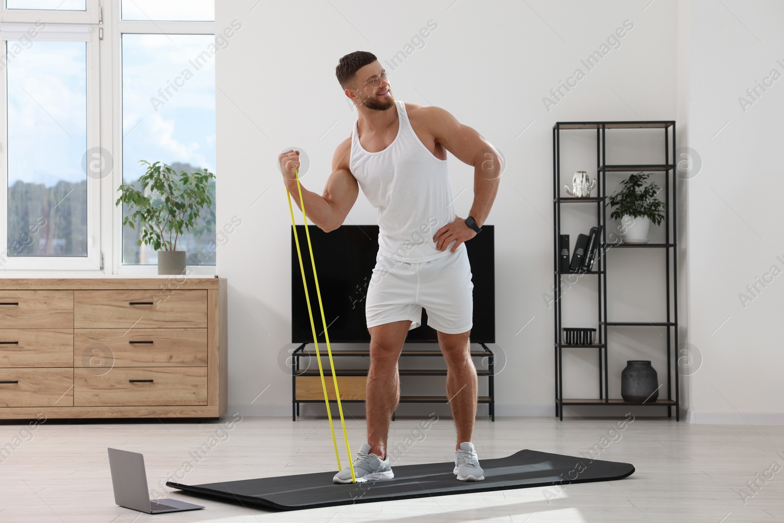 Photo of Muscular man doing exercise with elastic resistance band near laptop on mat at home