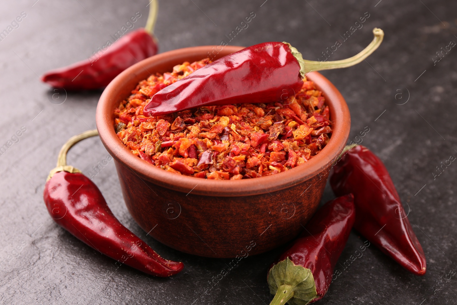 Photo of Chili pepper flakes in bowl and pods on dark textured table, closeup