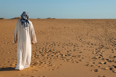 Man in arabic clothes walking through desert on sunny day, back view