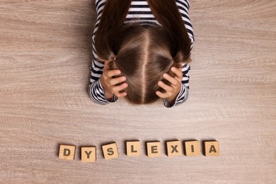 Little girl covering head with hands and cubes with word Dyslexia at wooden table, top view