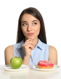 Doubtful woman choosing between apple and doughnut at table on white background