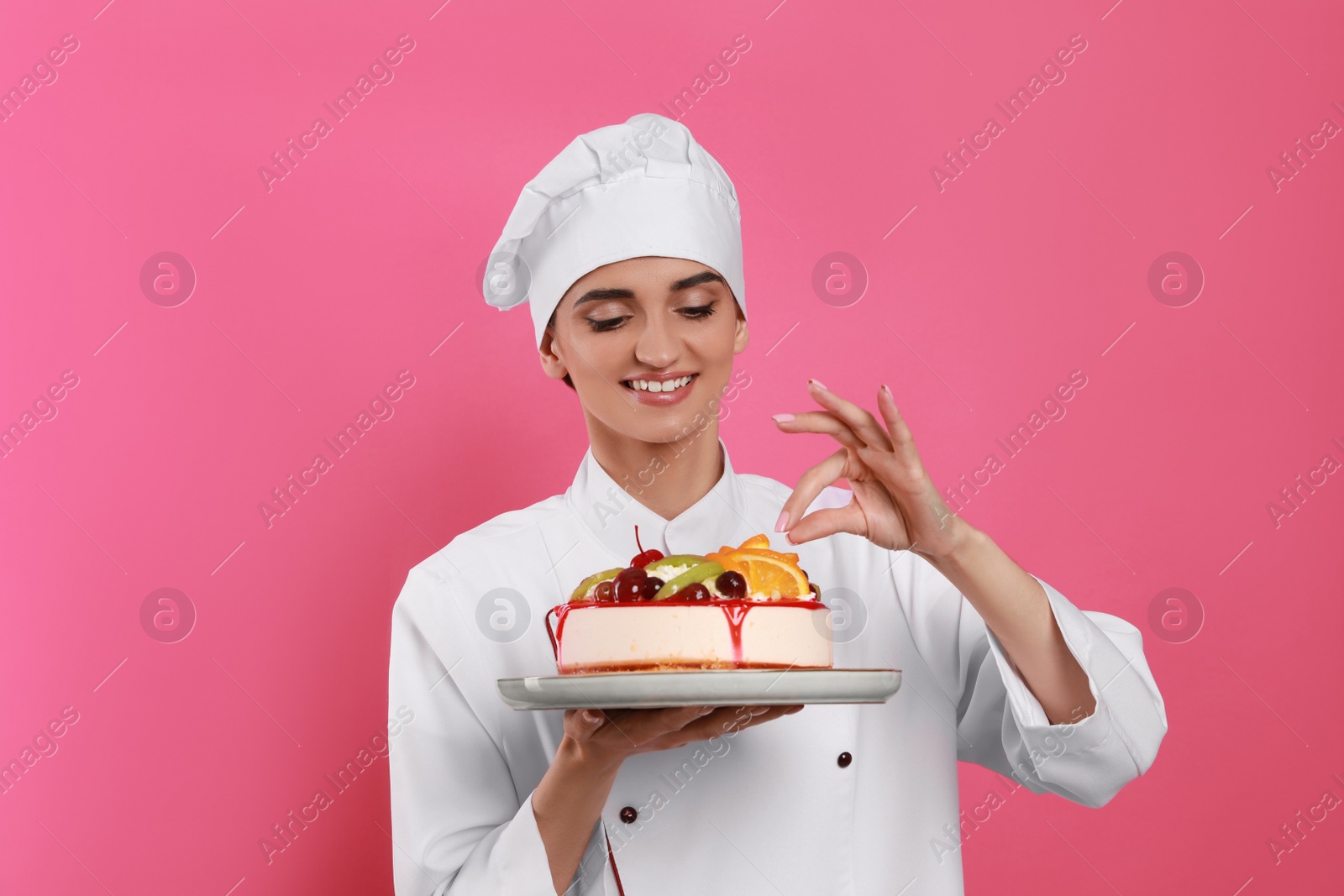 Photo of Happy professional confectioner in uniform decorating delicious cake on pink background