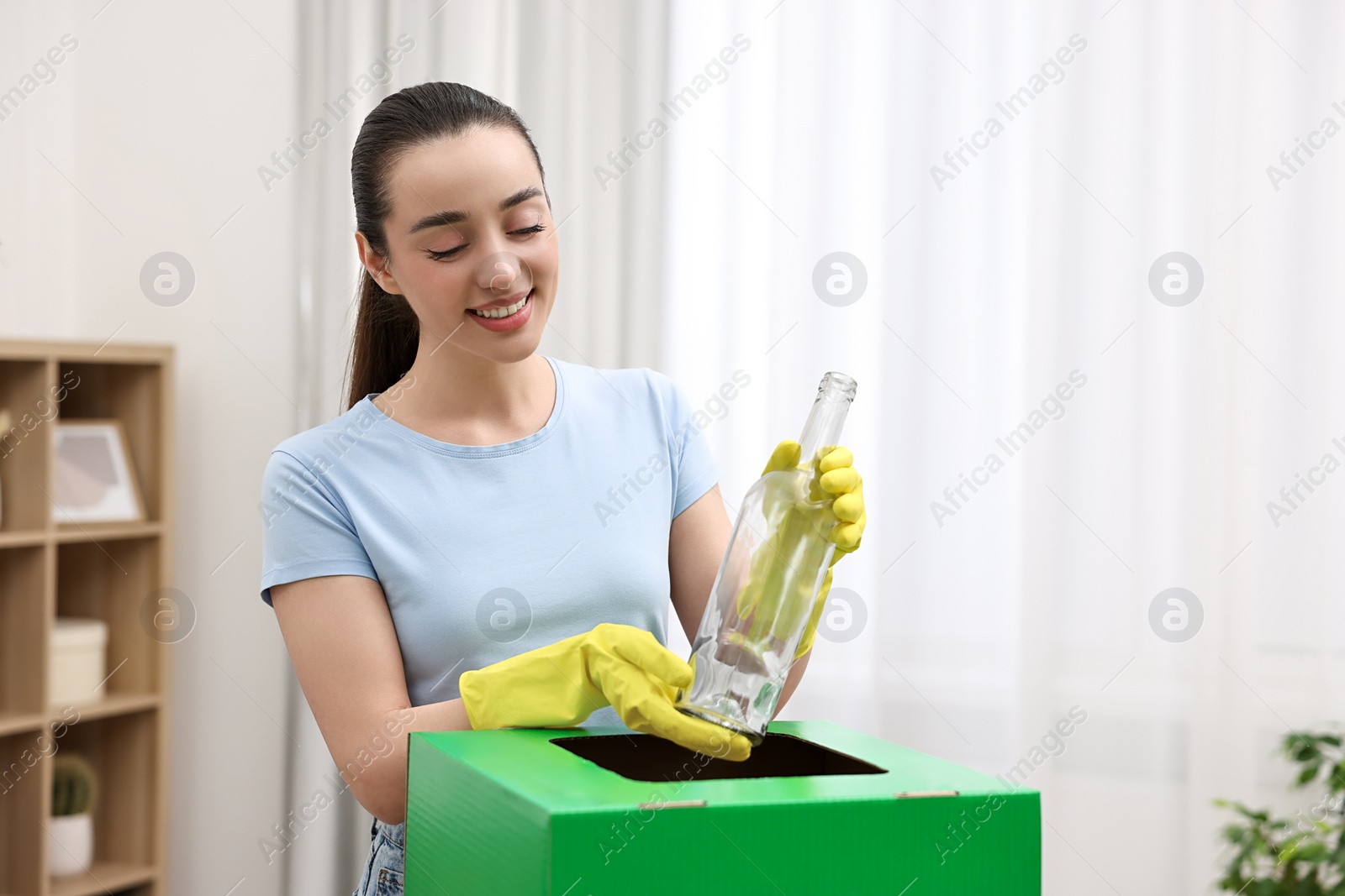 Photo of Garbage sorting. Smiling woman throwing glass bottle into cardboard box in room