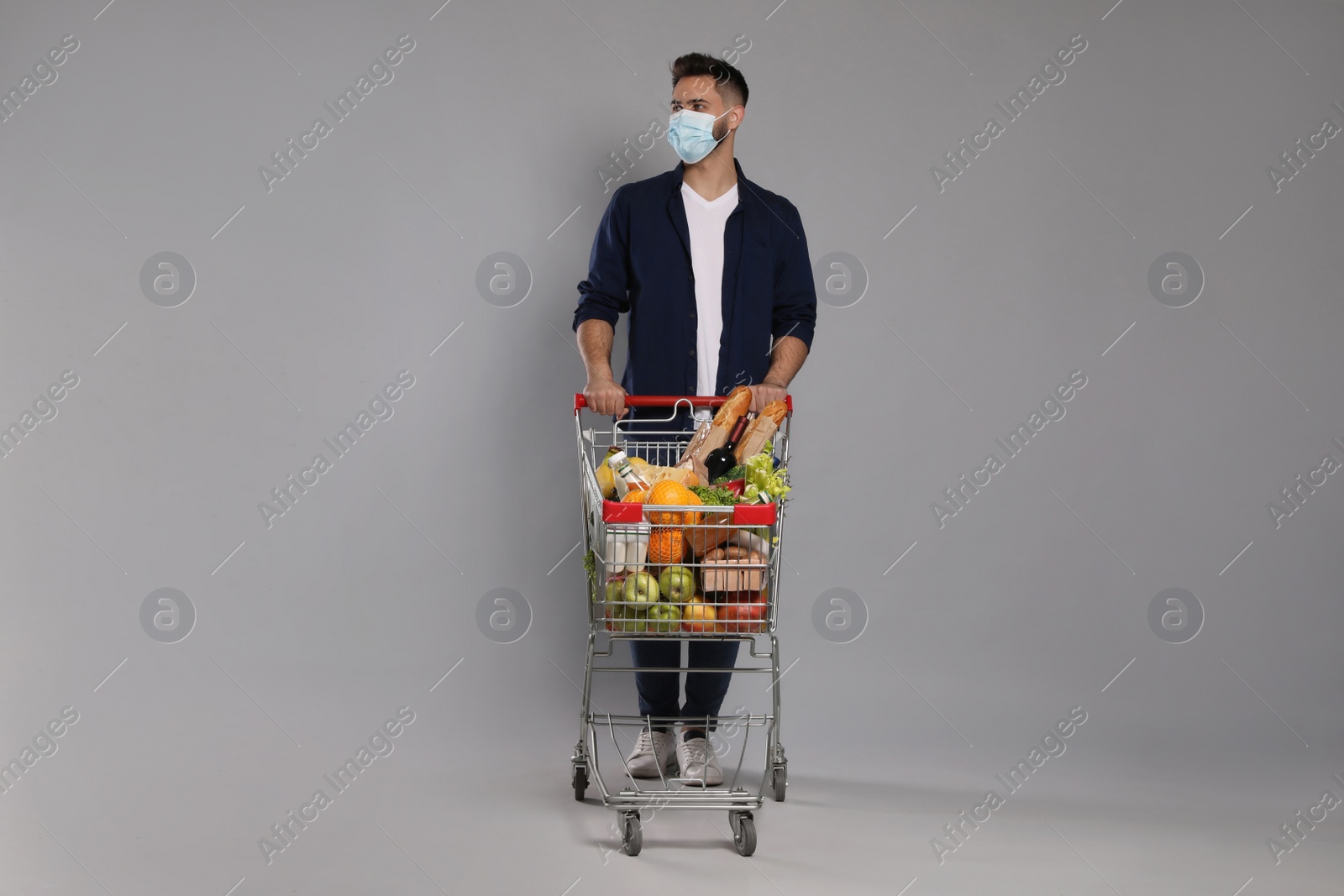 Photo of Man with protective mask and shopping cart full of groceries on light grey background