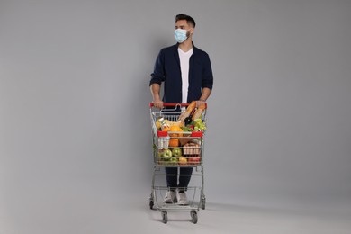 Man with protective mask and shopping cart full of groceries on light grey background