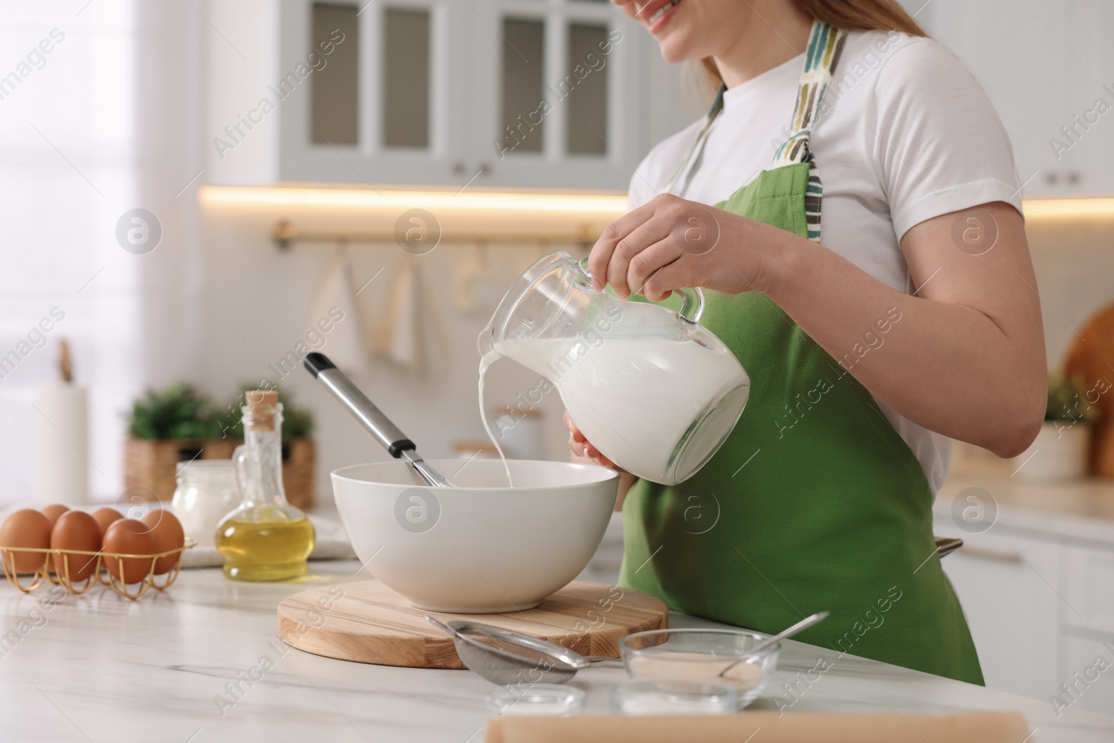 Photo of Making bread. Woman pouring milk into bowl at white table in kitchen, closeup