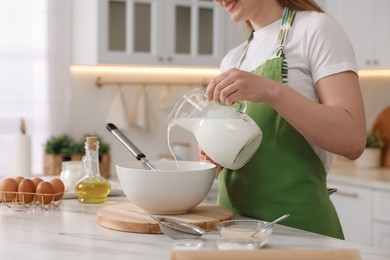 Making bread. Woman pouring milk into bowl at white table in kitchen, closeup