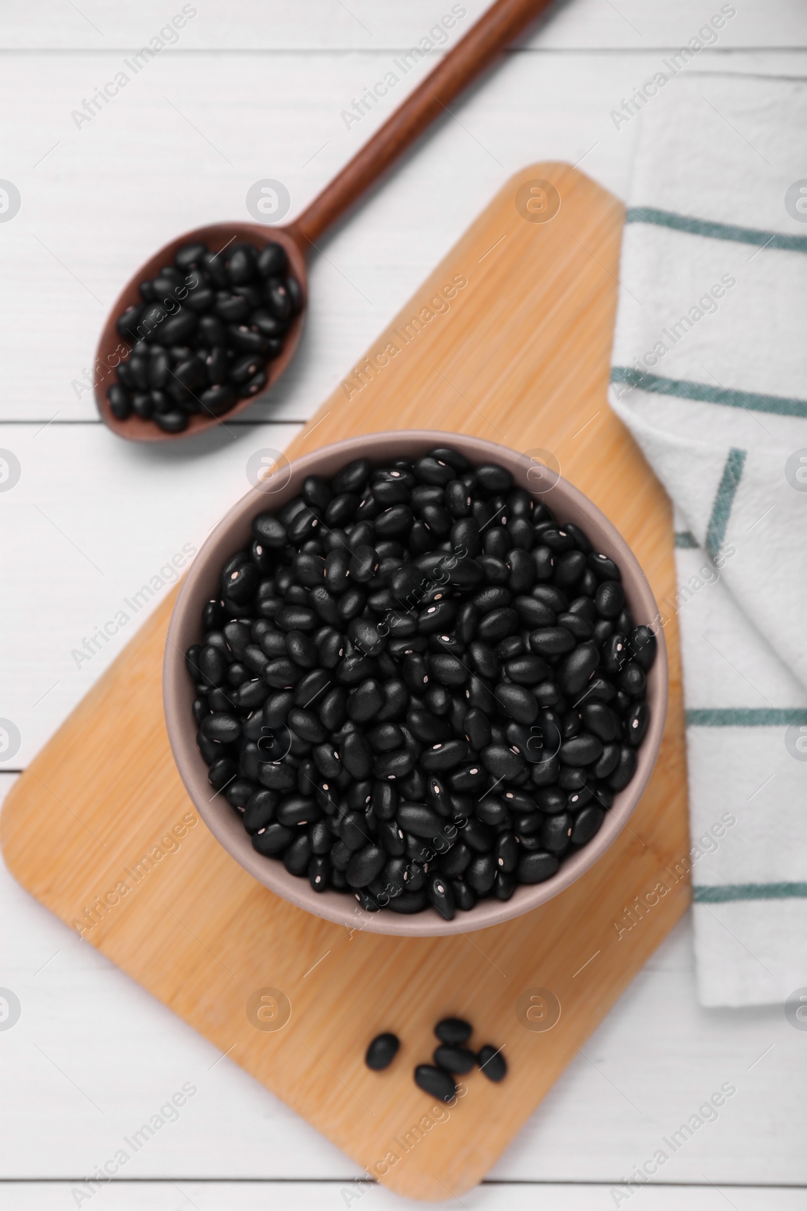 Photo of Bowl and spoon of raw black beans on white wooden table, flat lay