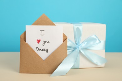 Photo of Happy Father's Day. Card with phrase I Love You, Daddy in envelope and gift box on beige table, closeup