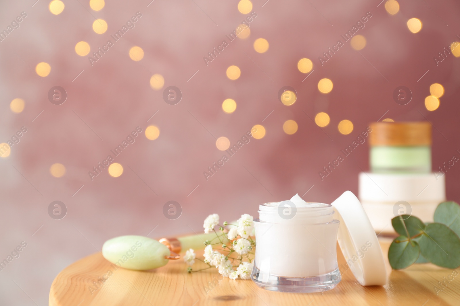 Photo of Natural face roller, cosmetic product and beautiful flowers on wooden table against blurred lights, space for text