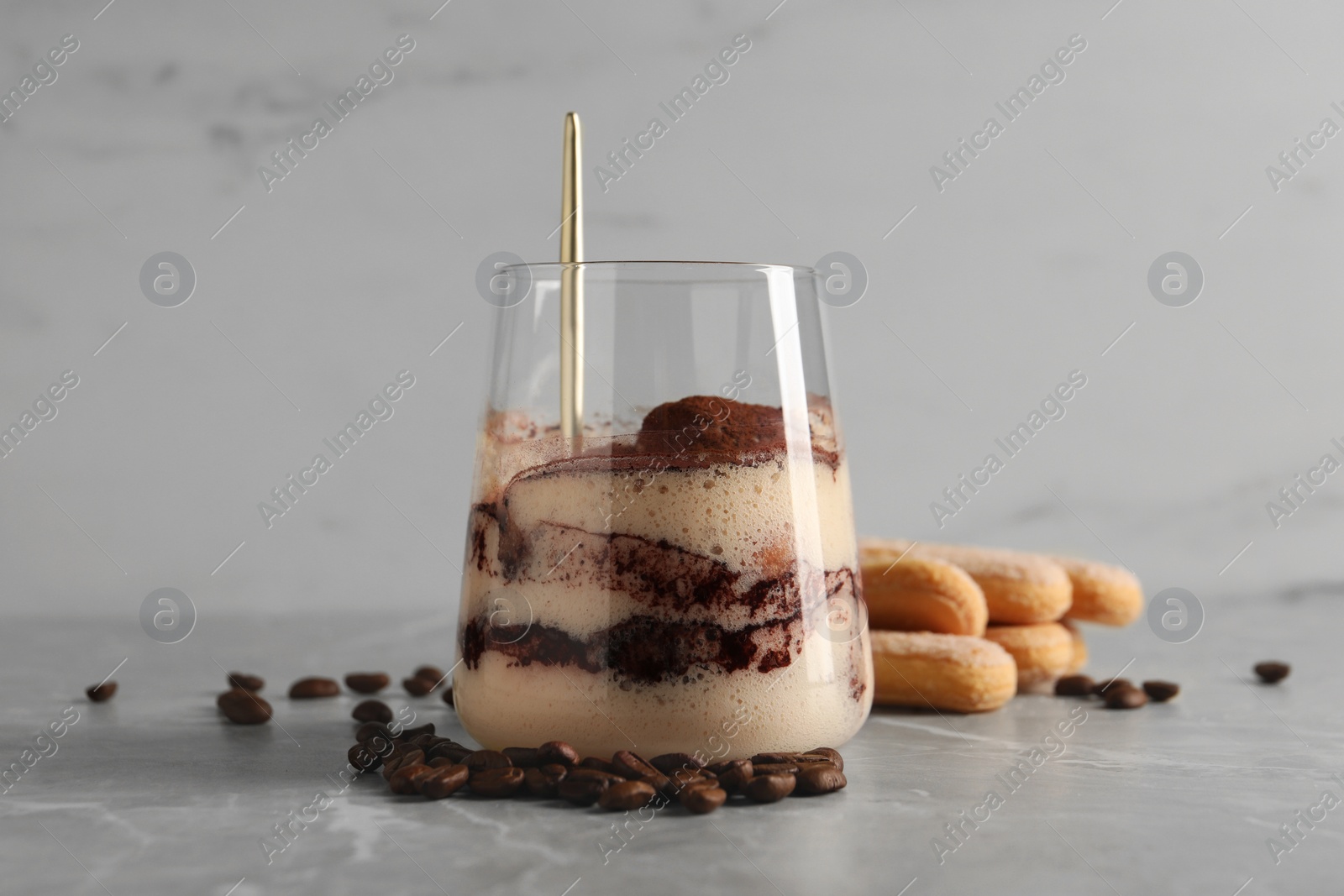 Photo of Tasty tiramisu in glass, coffee beans, spoon and biscuits on light grey table, closeup