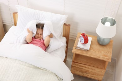 Photo of Sleepy girl covering ears with pillow near alarm clock at early morning indoors, above view