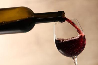 Pouring red wine into glass from bottle against blurred beige background, closeup