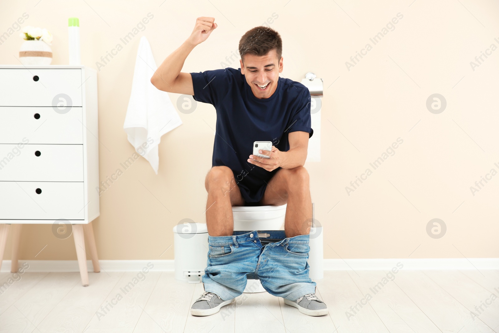 Photo of Young man using mobile phone while sitting on toilet bowl at home