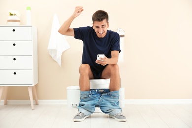 Young man using mobile phone while sitting on toilet bowl at home