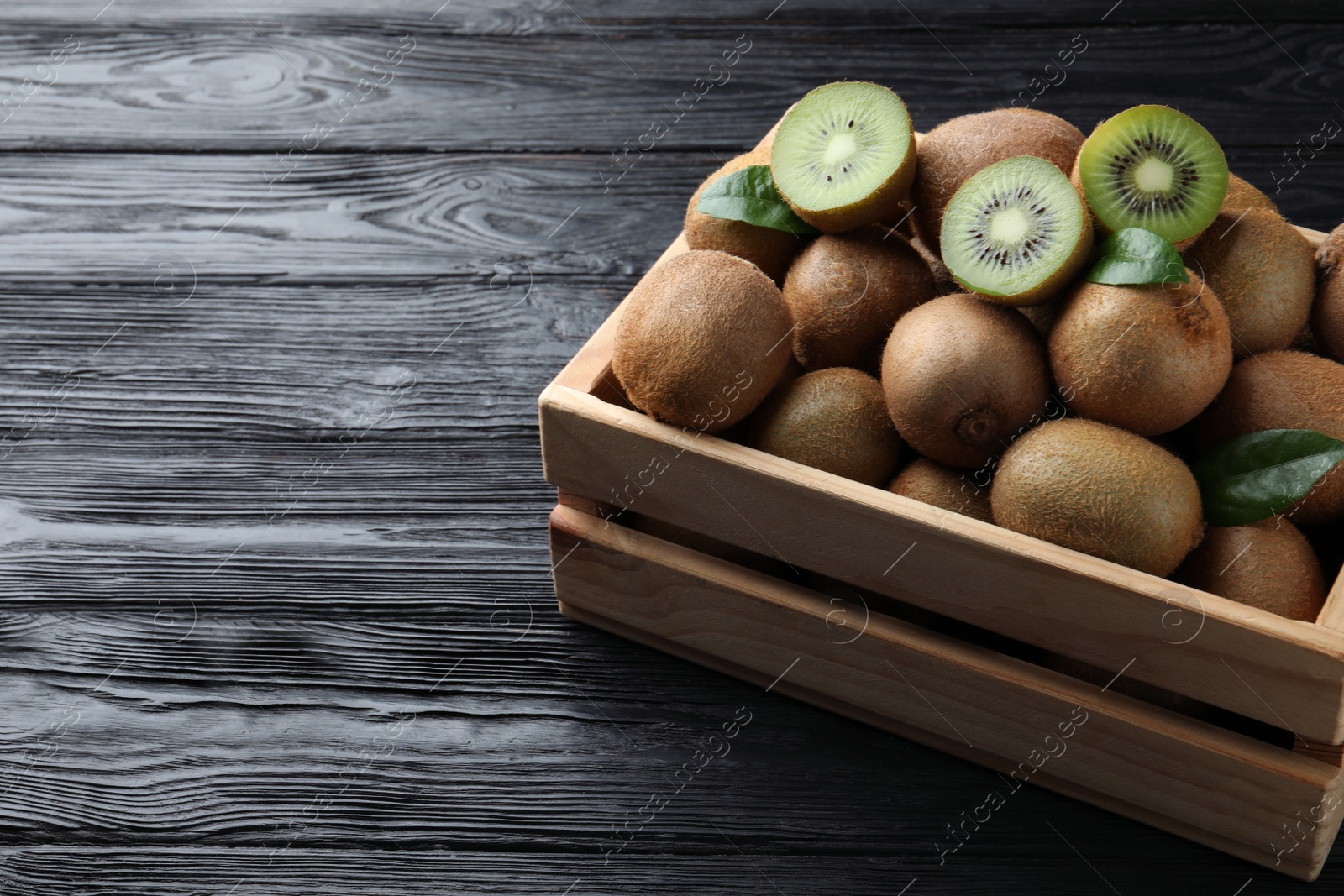 Photo of Crate with cut and whole fresh kiwis on black wooden table, space for text