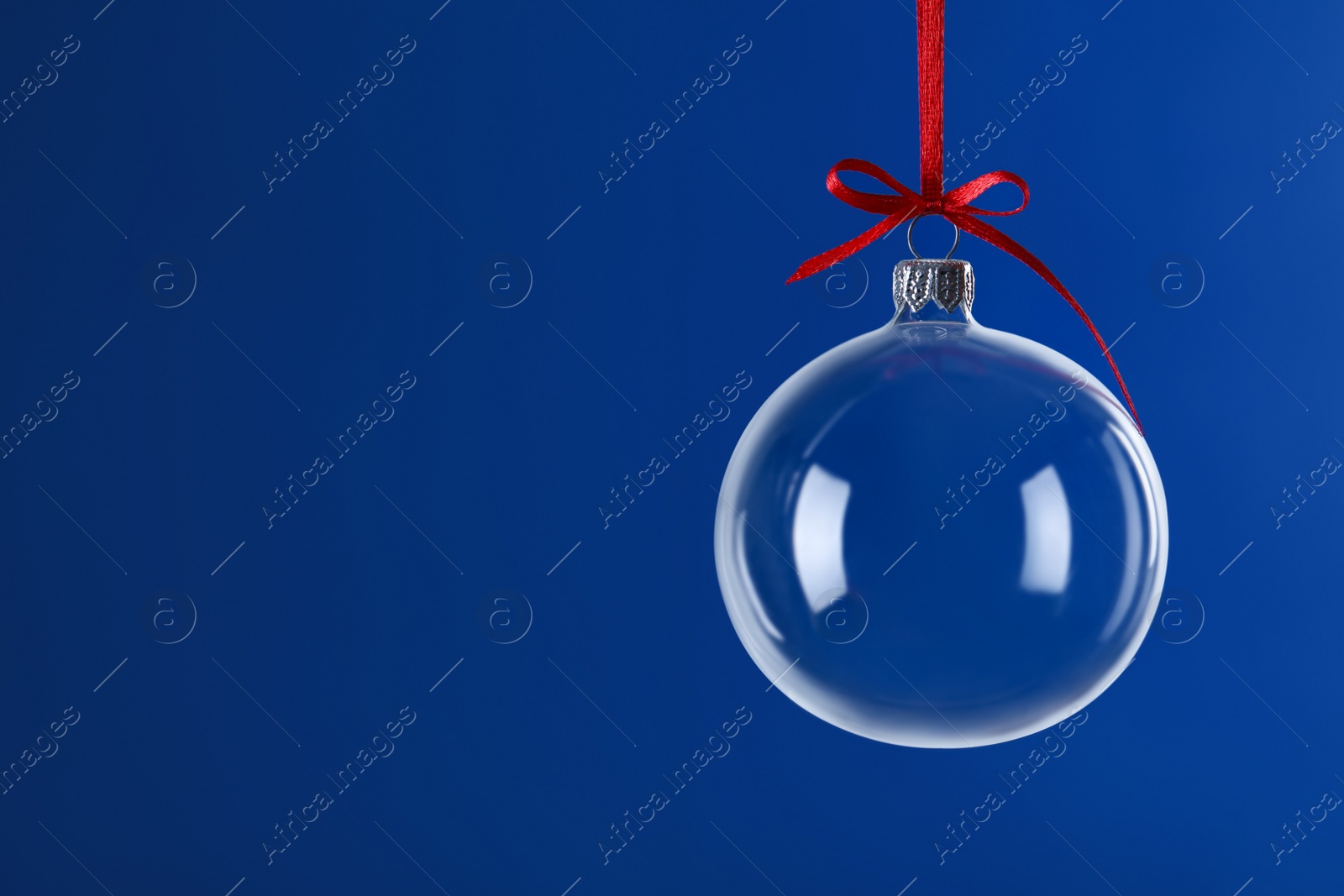 Photo of Transparent glass Christmas ball with red ribbon and bow against blue background. Space for text