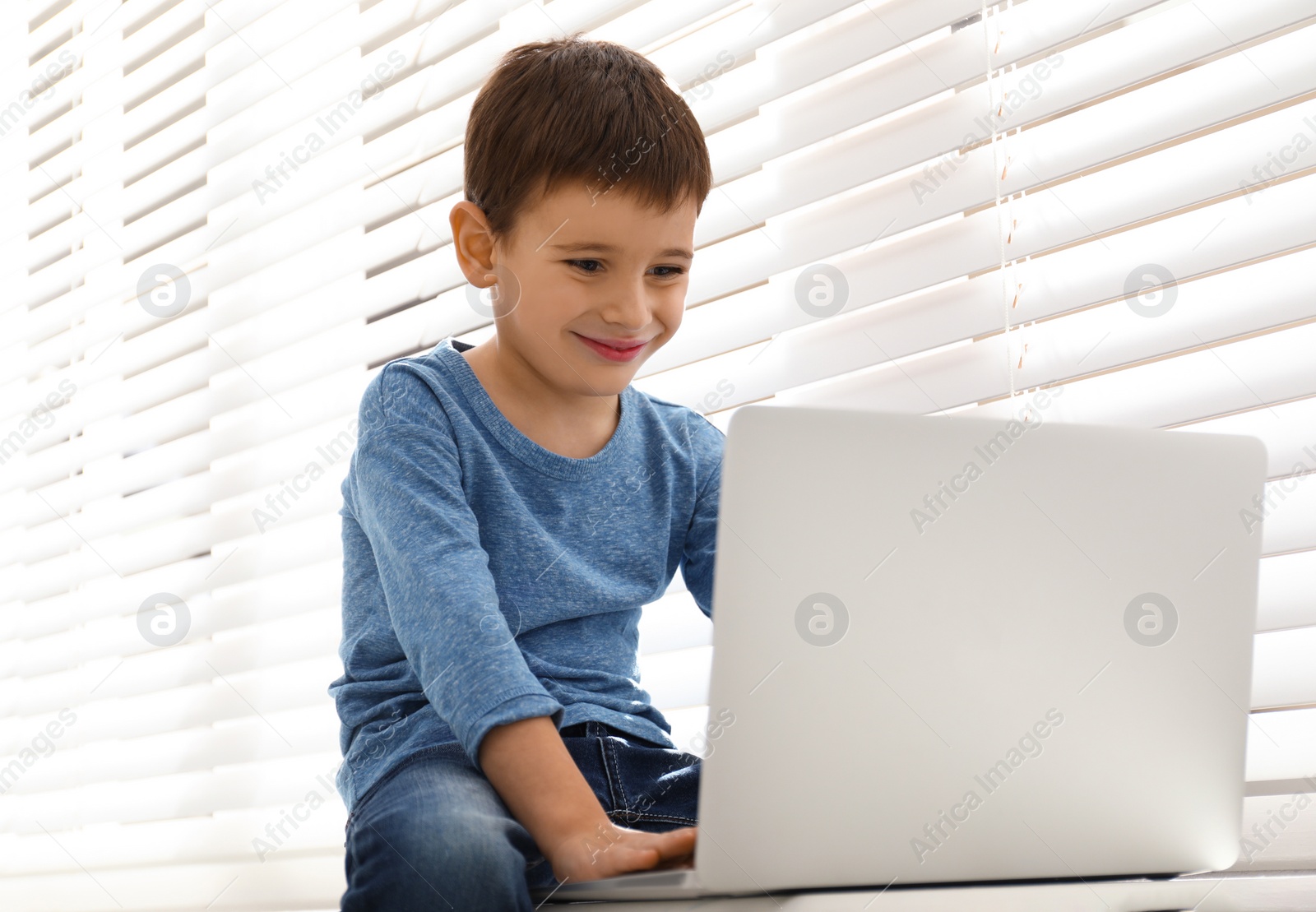 Photo of Happy little boy with modern laptop near window indoors