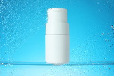 Photo of Bottle with moisturizing cream on light blue background, view through wet glass