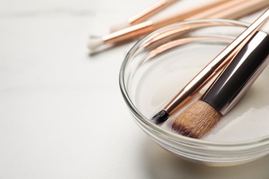 Clean makeup brushes in bowl with cleanser on white table, closeup. Space for text