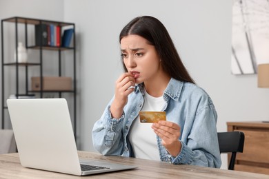 Photo of Stressed woman with credit card using laptop at table indoors. Be careful - fraud