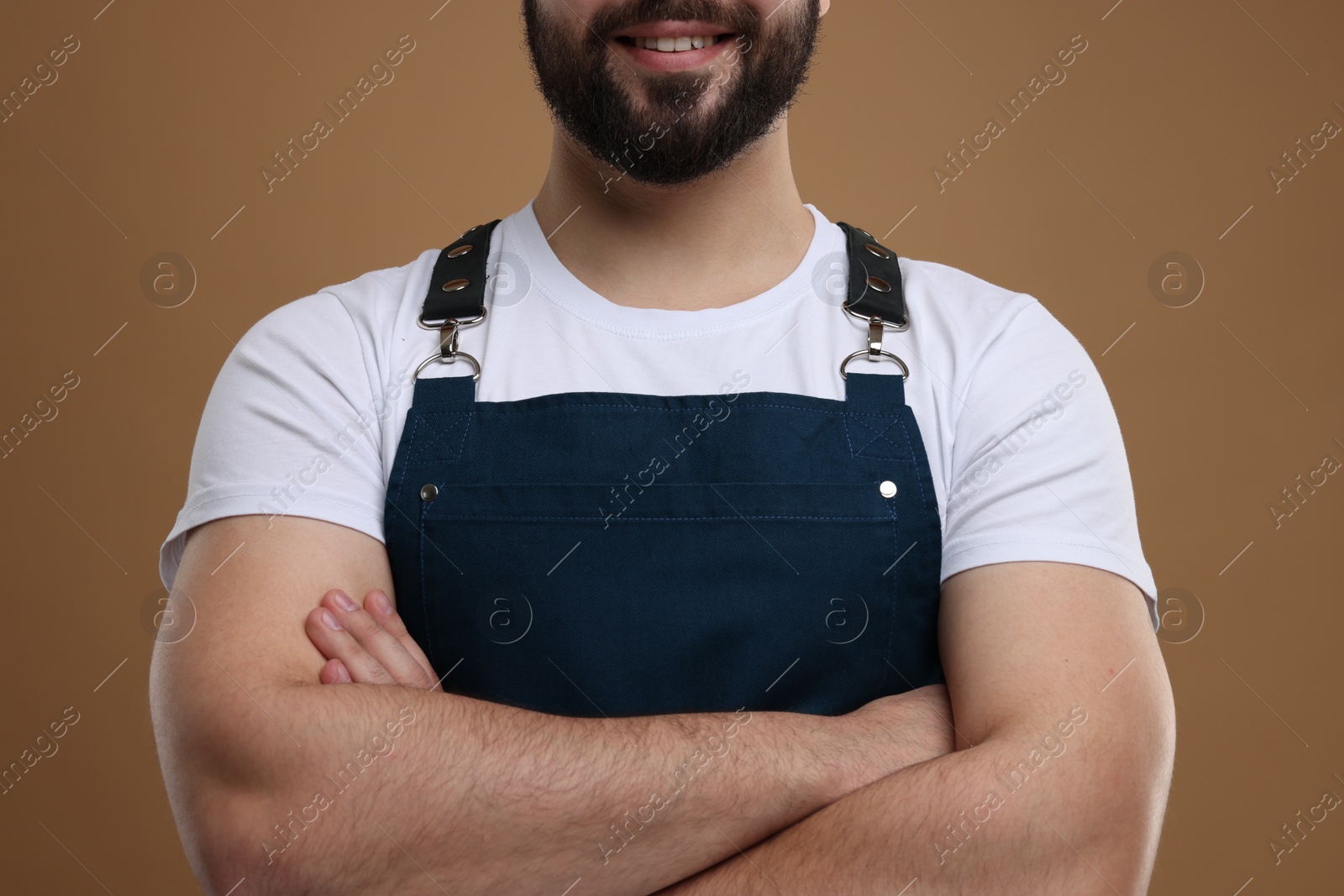 Photo of Smiling man in kitchen apron with crossed arms on brown background, closeup. Mockup for design