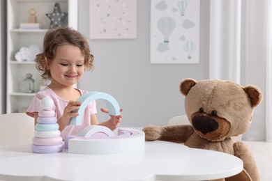 Cute little girl playing with toy and teddy bear at white table in room