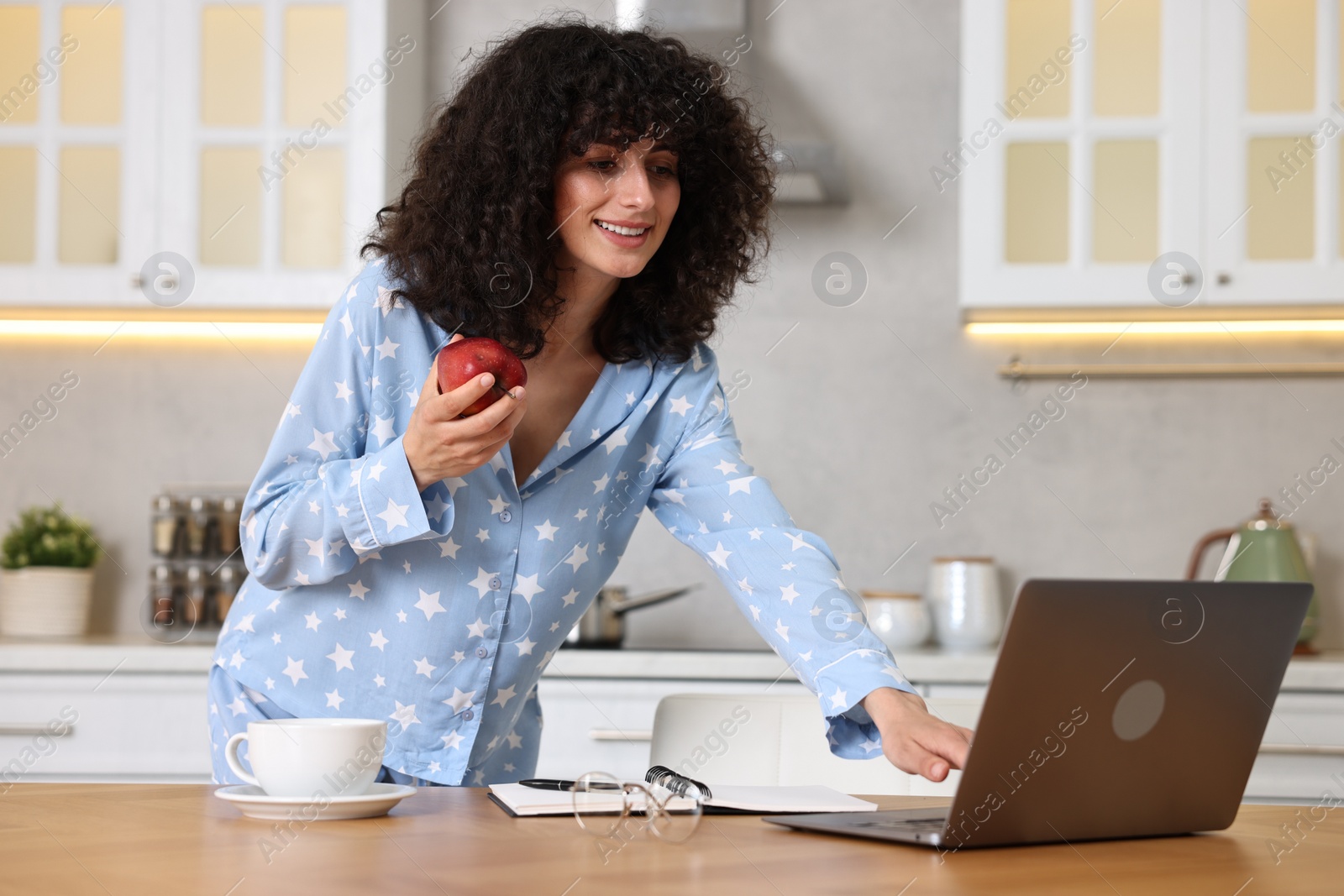 Photo of Beautiful young woman in stylish pyjama with apple using laptop at wooden table in kitchen