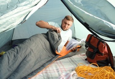 Photo of Young man reading book in sleeping bag inside of camping tent