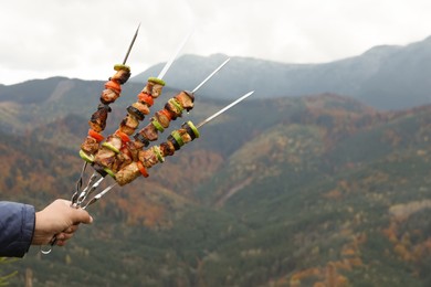 Photo of Man holding metal skewers with delicious meat and vegetables against mountain landscape, closeup