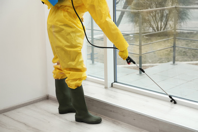 Photo of Pest control worker in protective suit spraying pesticide near window indoors, closeup