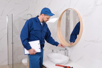 Professional plumber with clipboard checking water tap in bathroom