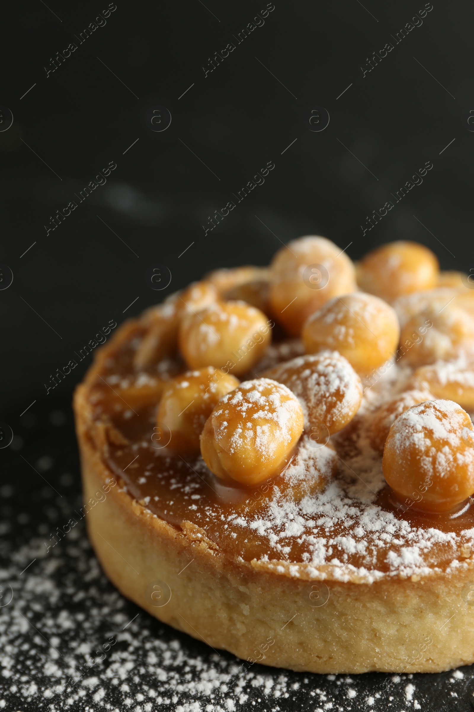 Photo of Delicious tart with hazelnuts, sweet caramel and powdered sugar on black table, closeup