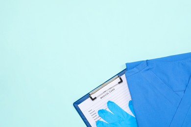 Medical uniform, clipboard and gloves on light blue background, flat lay. Space for text