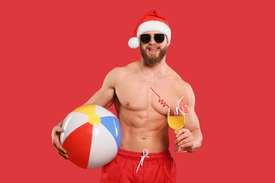 Muscular young man in Santa hat and sunglasses with ball and cocktail on red background