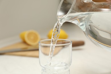 Pouring water from jug into glass on white table in kitchen, closeup