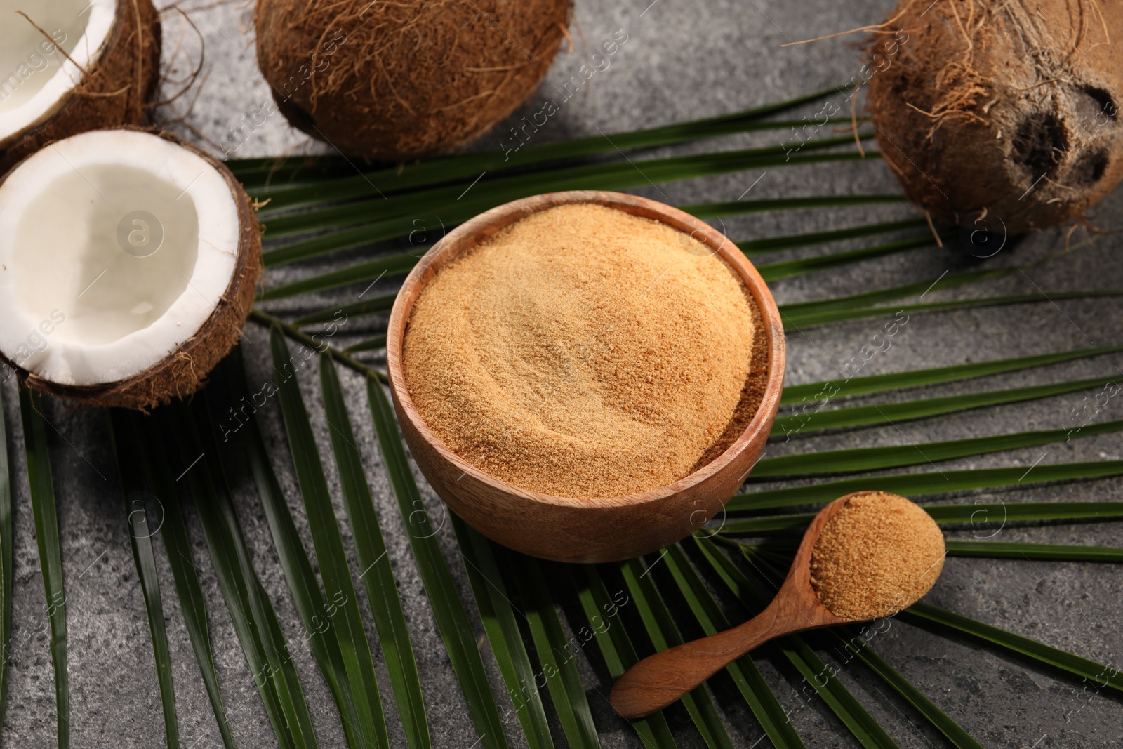 Photo of Coconut sugar in bowl, spoon, palm leaves and fruits on dark textured table, closeup